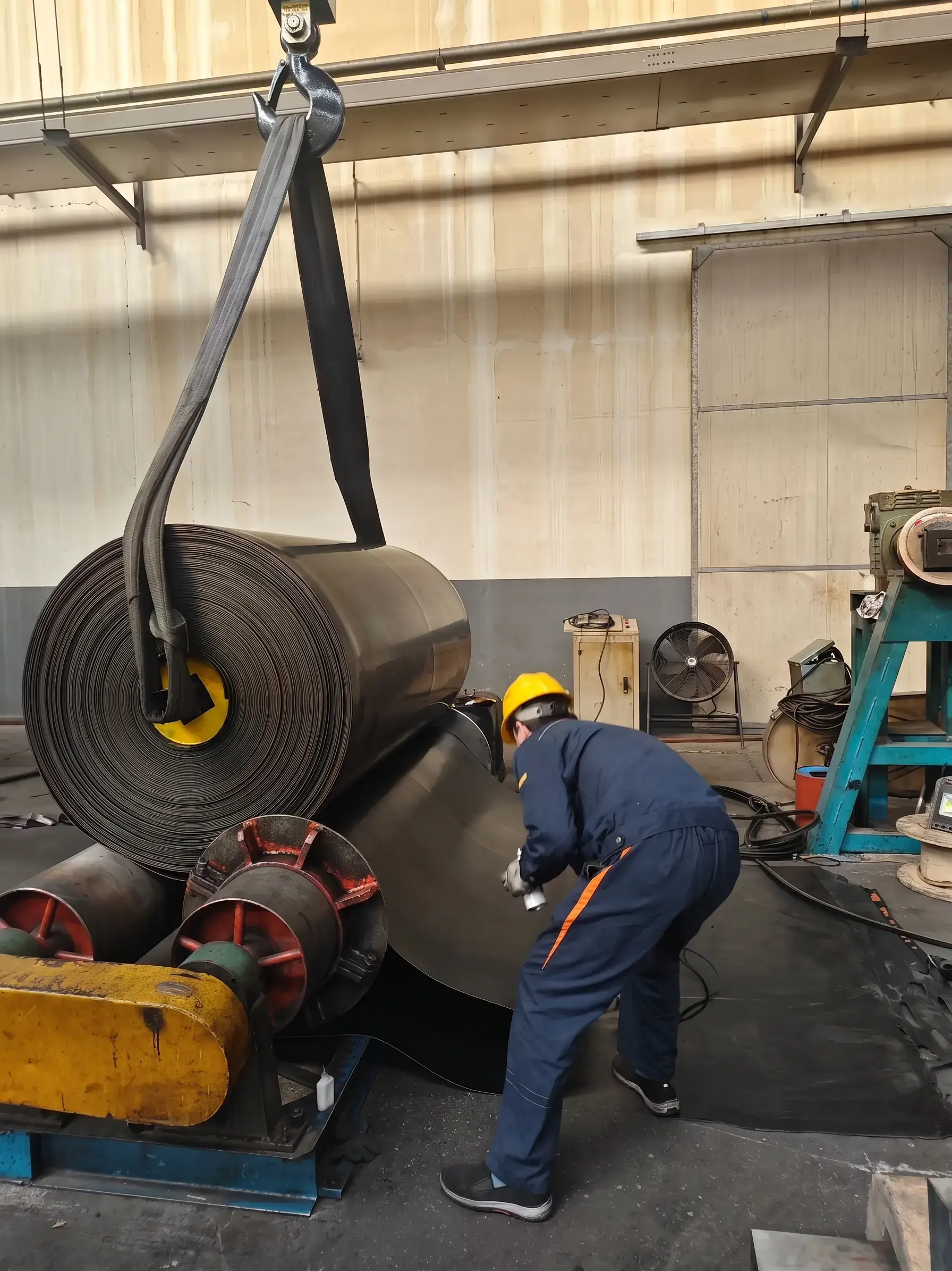 Factory worker lifting and handling heavy duty rubber conveyor belt roll using crane in manufacturing workshop